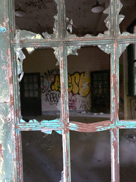 From outside of the Kings Park Psychiatric Center, looking through a broken multi-paned window at an empty room with two wooden doors visible in the back. Graffiti are painted on any free wall space.