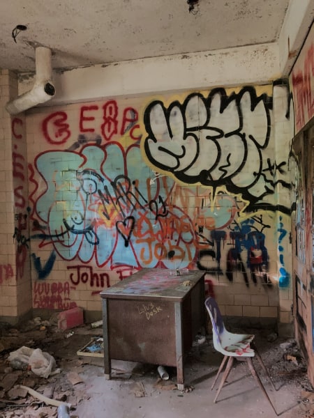 A small room in the Kings Park Psychiatric Center, with a desk and broken chair. The wall is full of graffiti, and there is litter and  debris on the floor, including what looks to be a plastic gas canister.