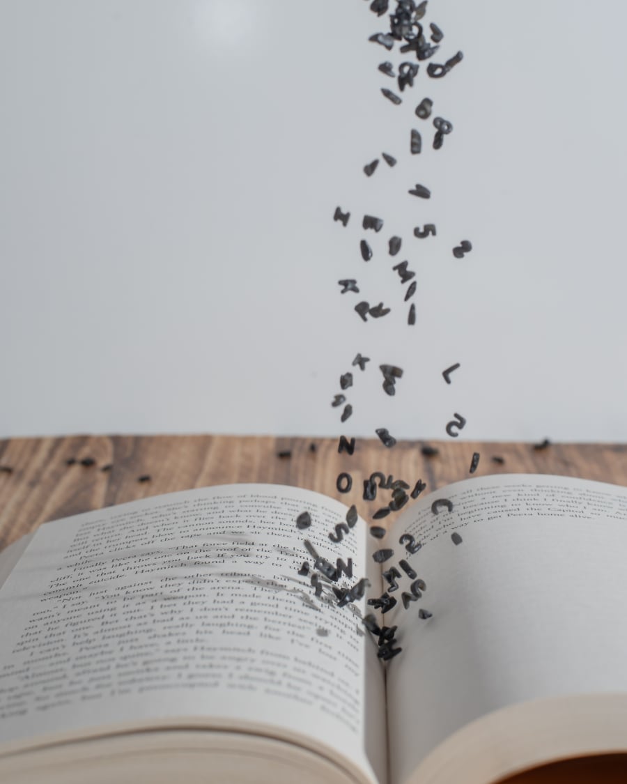 Photo composition by Racheal Halupa. Photo of an open hardcover book on a wooden tabel in front of a white background. There are small, black 3-dimensional English alphabetic letters falling from the top of the frame onto the crease of the open book.