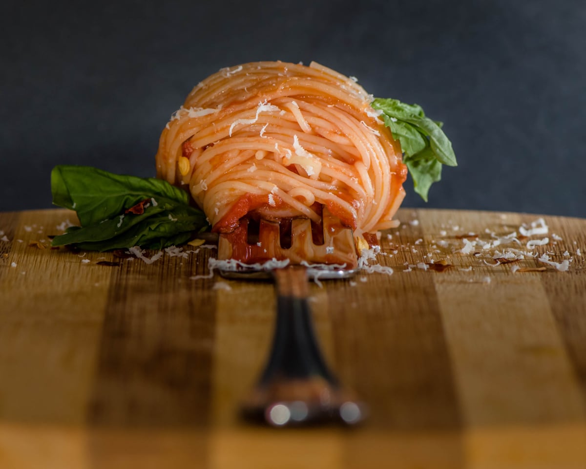 Photo by Racheal Halupa. A silver fork with tightly wound, lightly sauced spaghetti sits on a wooden cutting board. The fork is eye-level to the viewer and looks elongated in this perspective, with the bottom of the handle closest to the viewer and slightly blurred. The fork has some deeply green basil leaves attached to either side of the spaghetti. A light amount of paramasian cheese and red pepper flake has been sprinkled over the spaghetti.