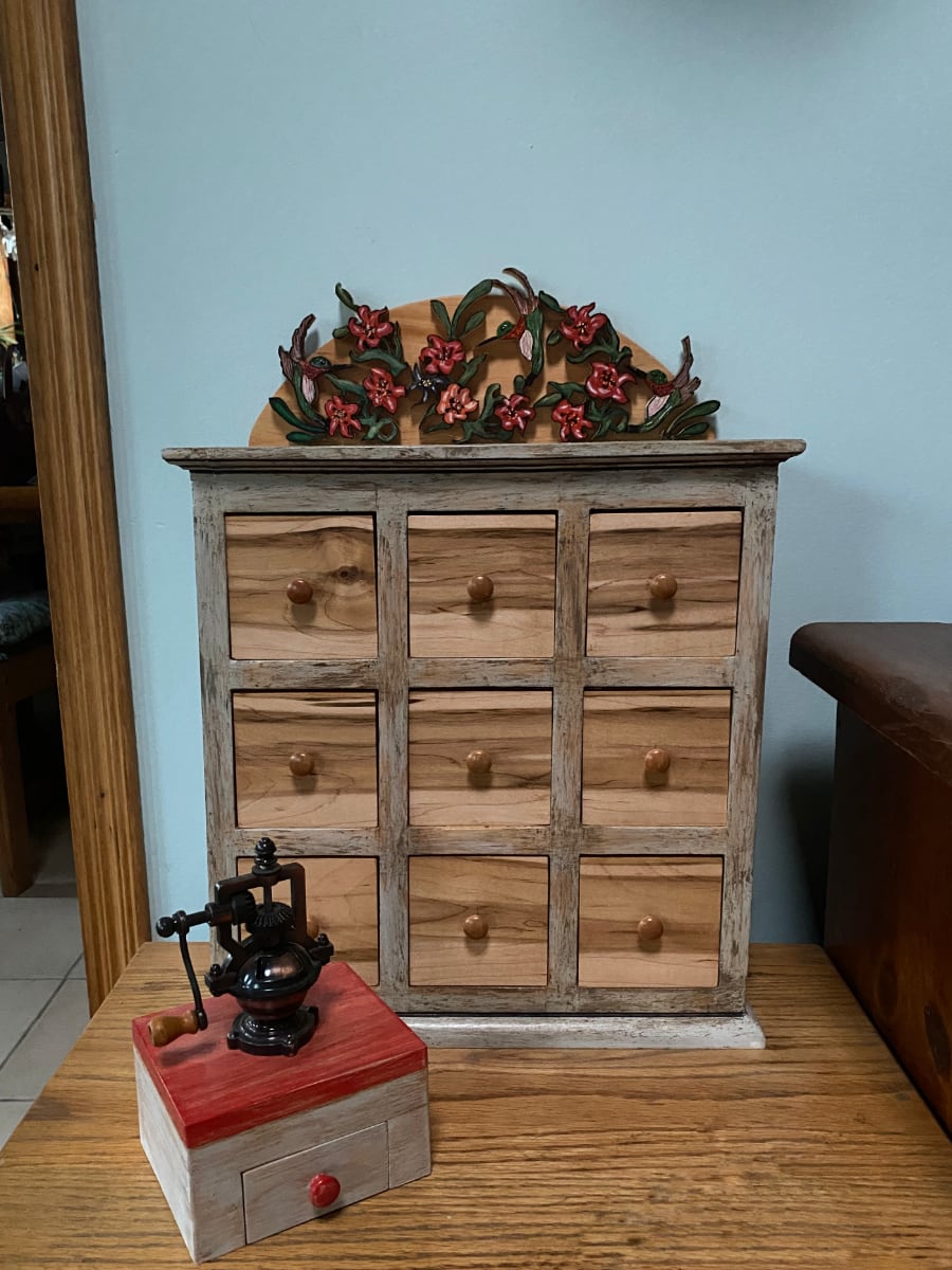 A photo of a wooden spice cabinet with drawers made of buthcer block wood and a small antique brass spice grinder on a wooden block with a small drawer to remove the ground spices. The top of the spice cabinet has a wooden design of red flowers and green vines. The wood top piece of the spice grider top is stained red. The remaining pieces of wood have a light whitewash over them. 