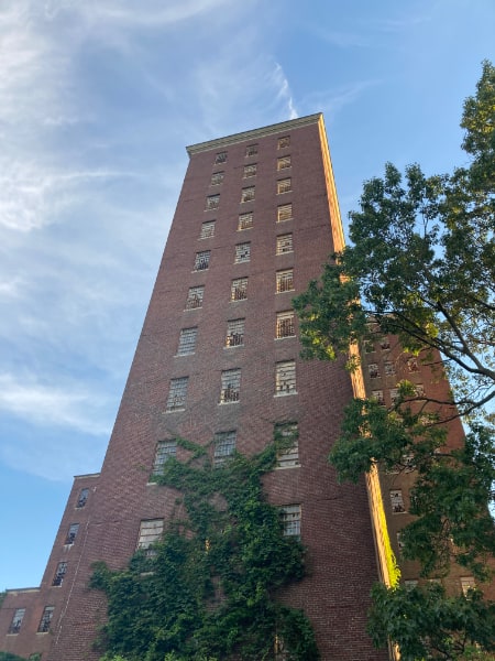 Building 93, a thirteen-story tower, at the Kings Park Psychiatric Center. Green vines crawl up the front, reaching to the fourth story. Many windows are broken. A partially visible leafy tree is visible on the right of the photo. It is a mostly clear day, with wispy clouds visible.
