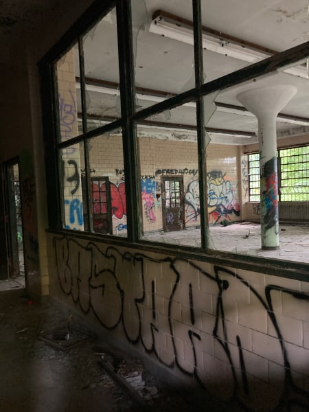 Inside the Kings Park Psychiatric Center, in a corridor where a large room with walls painted with graffiti is visible through window frames with broken glass. There is some litter strewn about in the corridor, but less than might be expected. The large room is cleared out, and the fluorescent fixtures on the ceiling are empty of light tubes.