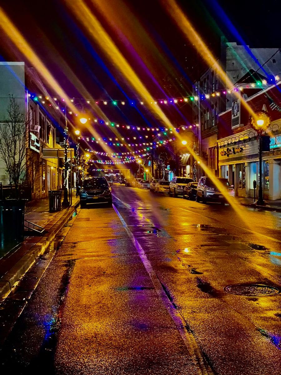 A saturated photo at night of Atlantic Ave, a northbound, one-way street, in the downtown of Lynbrook, New York, looking south from Merrick Rd. The street is wet from rain, though it is not raining in the photo. The street is empty, but there are cars parked on both sides of the road, facing the photographer. The road is decorated with colored lights going across the street from pole to pole and with white lights twisted around the poles. The road is lit with yellowish streetlights that are bright, and they leave the road bathed in golden light. The streetlights and the blue and green lights of the strands traversing the road are flaring, each with a beam extending from the upper left to the lower right with the light source in the center of the beam. There are parking meters, trash cans, and storefronts on either side of the road, with the names of a Hallmark store and a store called Card Shack readable on the right side of the photo.