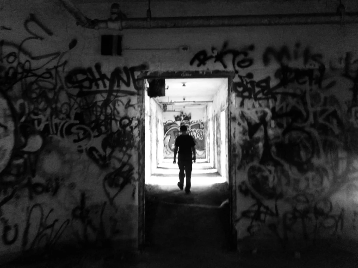 A black and white photo of a concrete wall, covered with graffiti, water or fire-extinguisher pipes by the ceiling, in shadow. In the lower middle of the wall is a doorway into a hallway that is well-lit. There is a young man in silhouette walking away from the viewer. There appears to be several doorways off the corridor. The end of the hallway is another wall covered with graffiti.