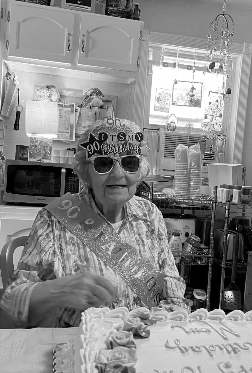 A black and white photograph of a 90-year old woman celebrating her birthday. She wears a tiara, sunglasses, and a sash. The tiara is topped with the numbers "90." The glasses have words attached to the top of them, "90 It's My Birthday" And the sash says "90 and Fabulous." There is a cake partially visible in the lower right of the photo. The background of the photo is a kitchen -- a microwave oven and an air conditioning unit are visible, along with sundry pantry items. The woman — a mother, grandmother, and great-grandmother — is smiling.