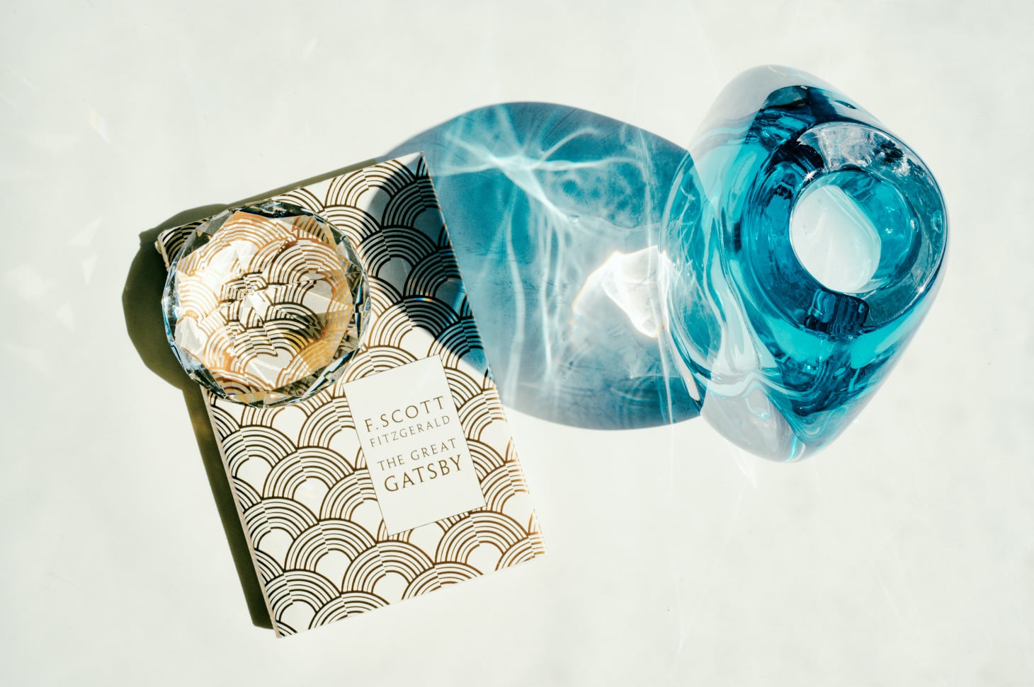 A copy of The Great Gatsby on a white background with a translucent blue vase and a polyhedral clear crystal.