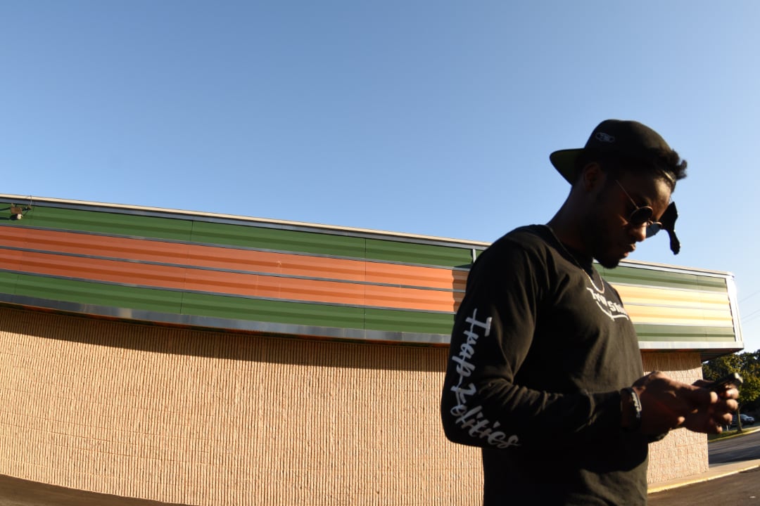 A man standing in a parking lot. He wears sunglasses, a baseball cap, and a pick in his hair. He is standing in front of a building that has a green and orange striped awning on its facade.